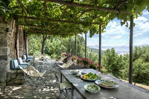 An outdoor dining area with stone tables and chairs, surrounded by lush greenery and a view of distant hills.
