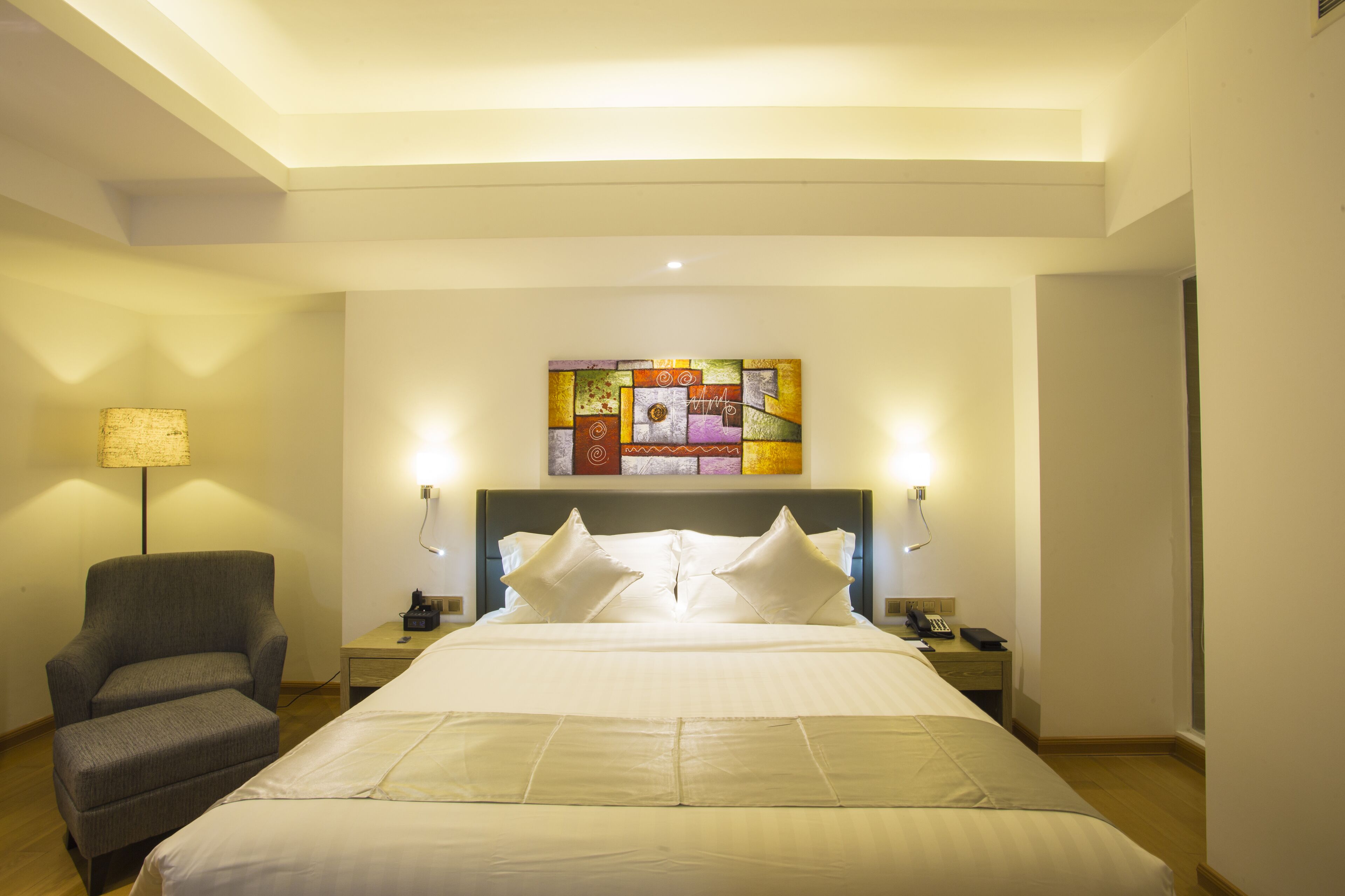 A hotel room with a large bed, a chair, a bedside table with a lamp, and a wall art piece.