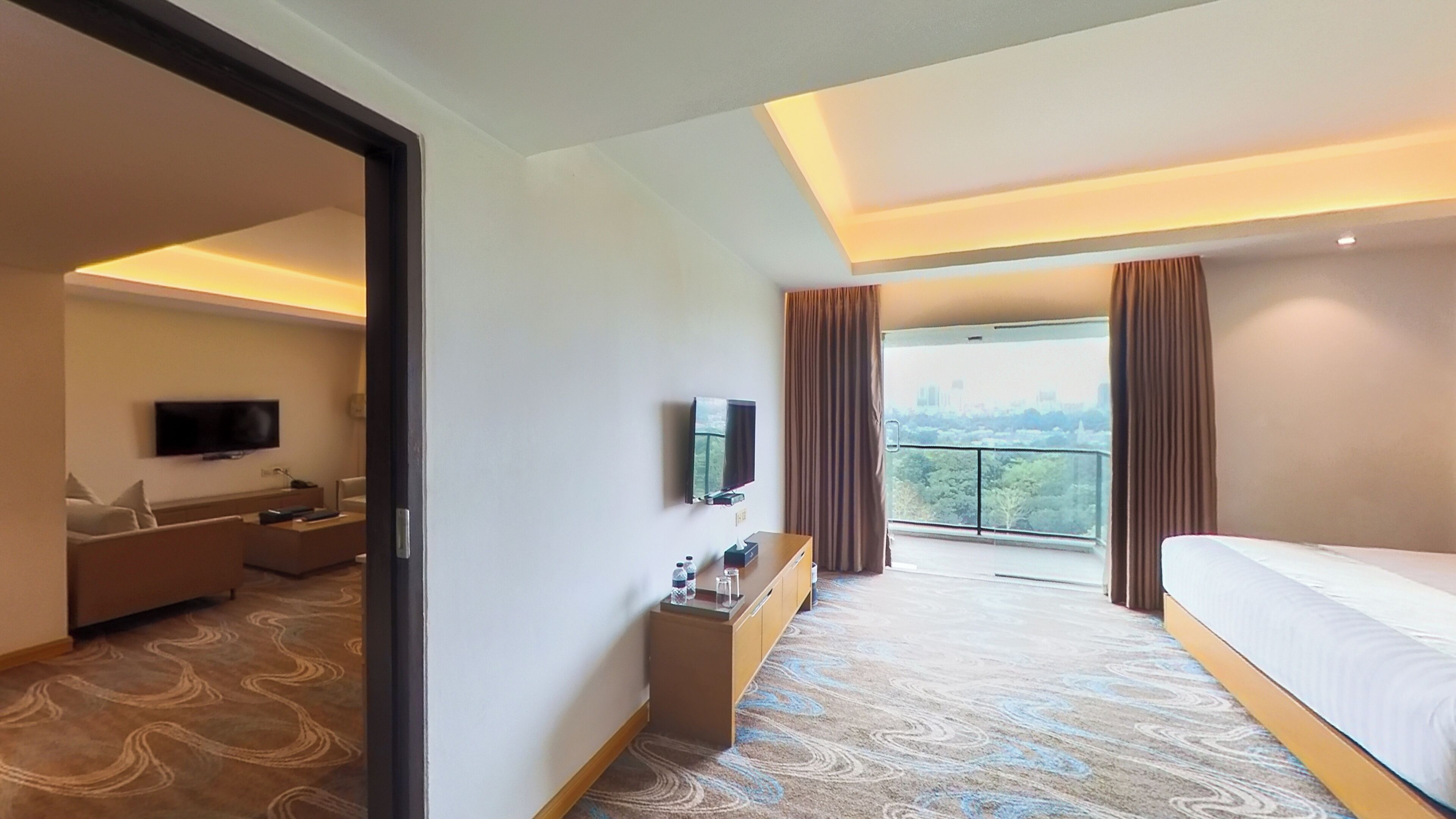 Habitación de hotel con cama, televisor, escritorio, silla, una mesita con una botella y vistas a un paisaje urbano a través de una ventana.