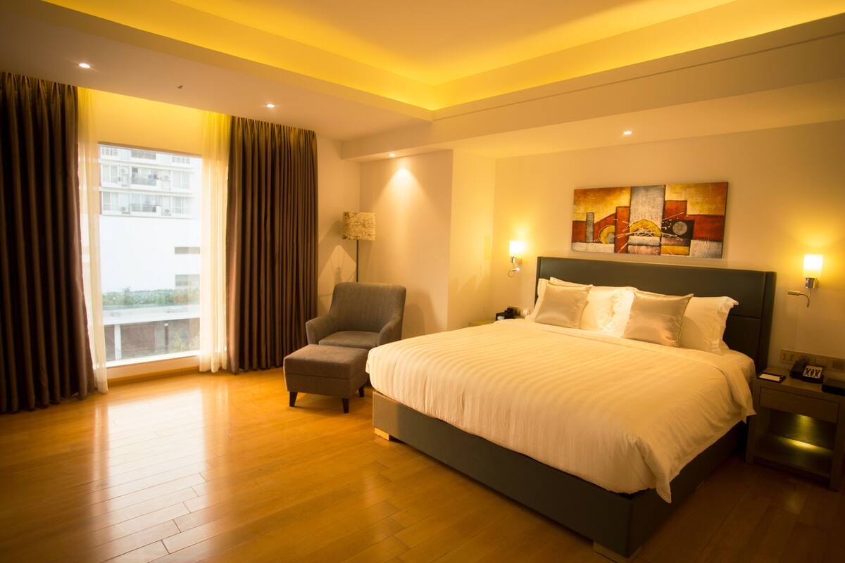 A hotel room with a large bed, a chair, a nightstand, a lamp, and a painting on the wall.