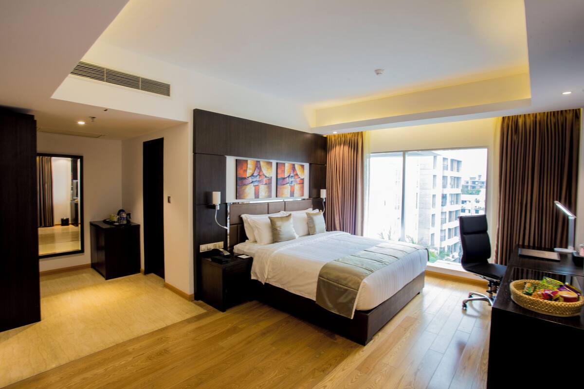 A modern hotel room with a large bed, a desk, a chair, a desk lamp, and a window with curtains.