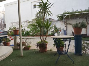 Un espace extérieur aménagé avec des tables et des chaises, entouré de plantes en pot et avec un bâtiment blanc en arrière-plan.