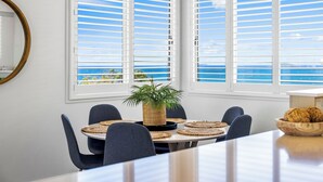 Comedor con una mesa dispuesta para seis personas, rodeada de sillas, y vista al océano a través de amplias ventanas con contraventanas blancas.