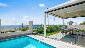 Una piscina exterior moderna con vistas despejadas al océano y un patio con mobiliario exterior.