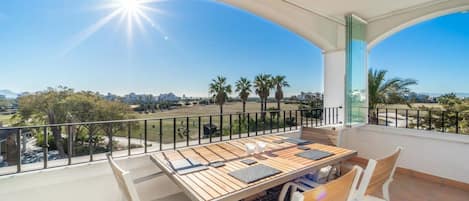 A balcony with a table and chairs, offering a view of a green landscape and buildings in the distance.