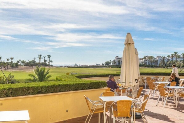 Ein Außenbereich mit Tischen und Stühlen, einem großen Sonnenschirm und Blick auf einen Golfplatz.
