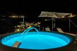 Une piscine agrémentée d’un élément décoratif aquatique, illuminée la nuit, entourée d’une clôture, et offrant une vue sur un paysage éclairé en arrière-plan.