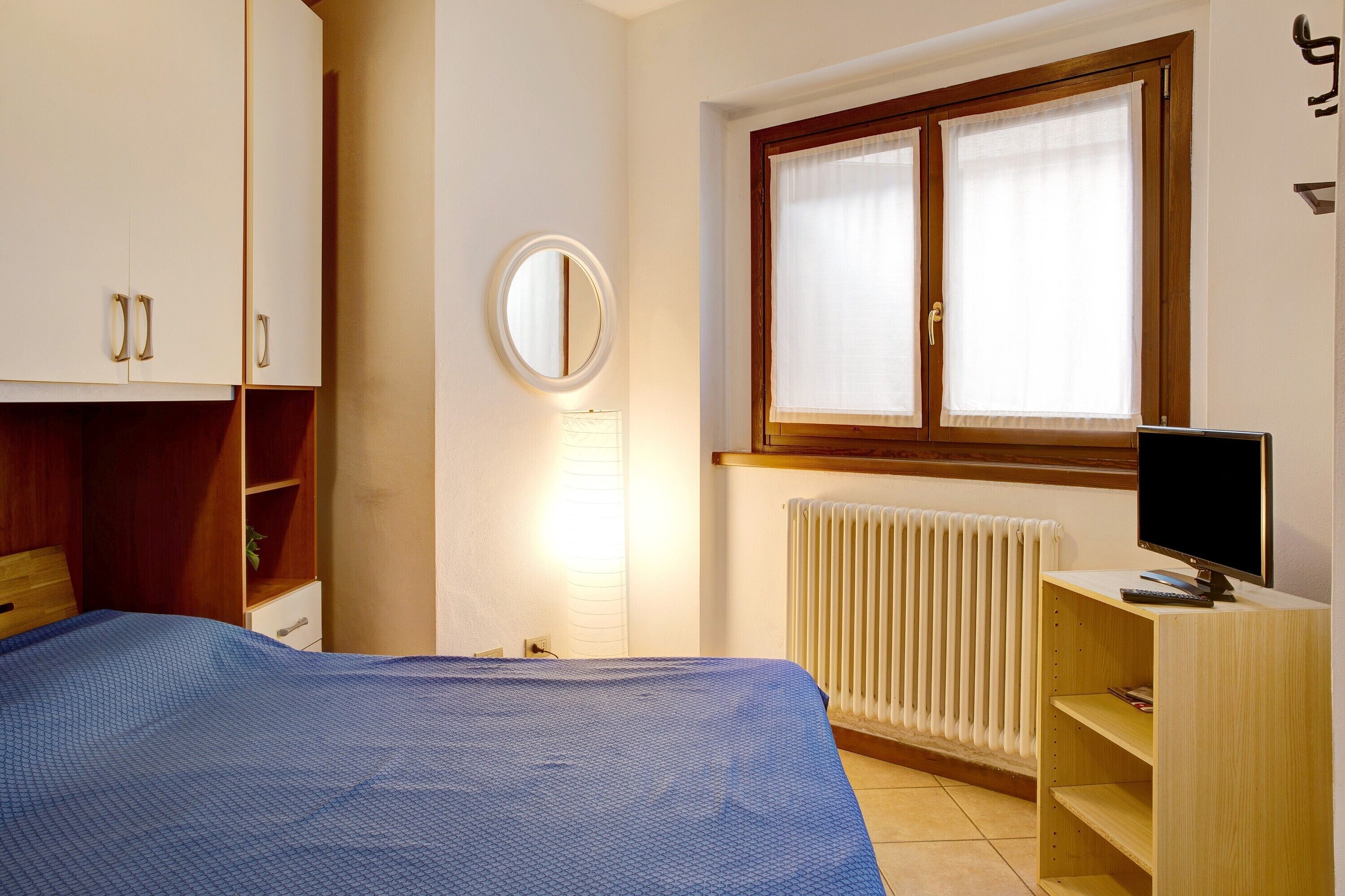 A bedroom with a bed, a bedside table with a TV, a window with curtains, a radiator, and a wardrobe.