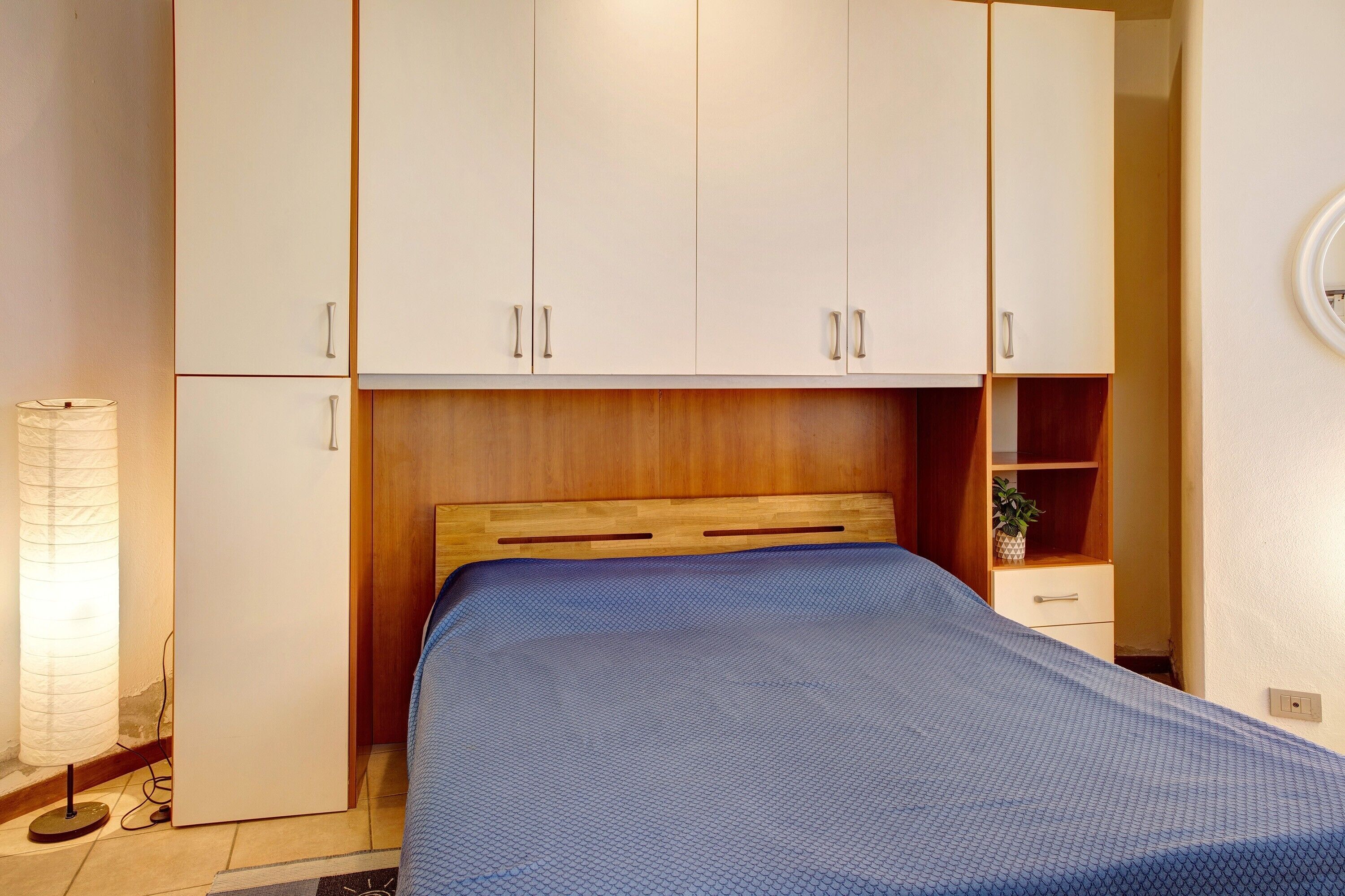 Un dormitorio con cama, cabecero de madera, armario blanco, mesita de noche y una lámpara.