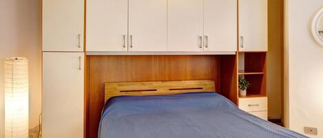 Un dormitorio con cama, cabecero de madera, armario blanco, mesita de noche y una lámpara.