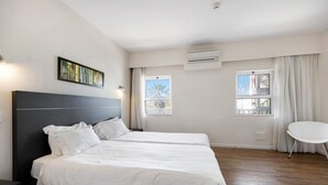 Una habitación de hotel con una cama grande, dos ventanas, un aire acondicionado y una mesita pequeña.
