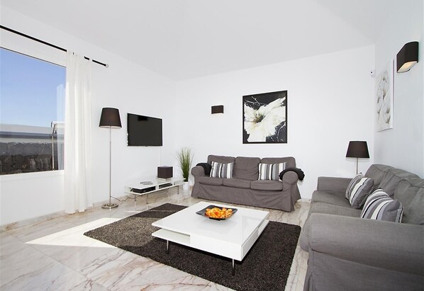 A modern living room with a grey sofa, a white coffee table, and a flat-screen TV mounted on the wall.