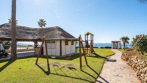 Um parquinho à beira-mar com baloiços, um pequeno edifício e um caminho de pedra.