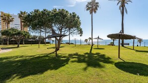 Uma área gramada ao ar livre com árvores e guarda-sóis de palha, céu claro e vista distante do mar.