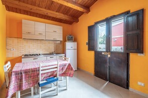 Een kleine keuken met houten kastjes, een kleine koelkast, een tafel met een geruit tafelkleed, en een raam met een donkere deur.