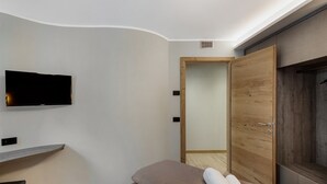 A modern hotel room with a flat-screen TV mounted on the wall, a wooden wardrobe, a bed with a brown headboard, and a small table with a chair.