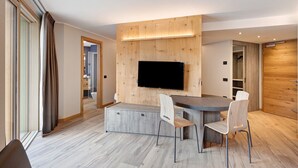 Ein modernes Wohnzimmer mit Flachbildfernseher an einer Holzwand, ein Essbereich mit Holztisch und -stühlen sowie ein Flur, der zum Badezimmer führt.