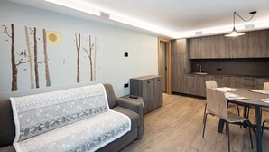 Un salon moderne comprenant un canapé, une table à manger et des chaises, un coin cuisine et un mur orné d’une fresque sur le thème de la nature.