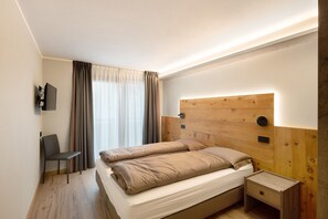 Une chambre avec une tête de lit en bois, un lit simple avec une literie beige, une table de chevet, une chaise et un téléviseur fixé au mur.