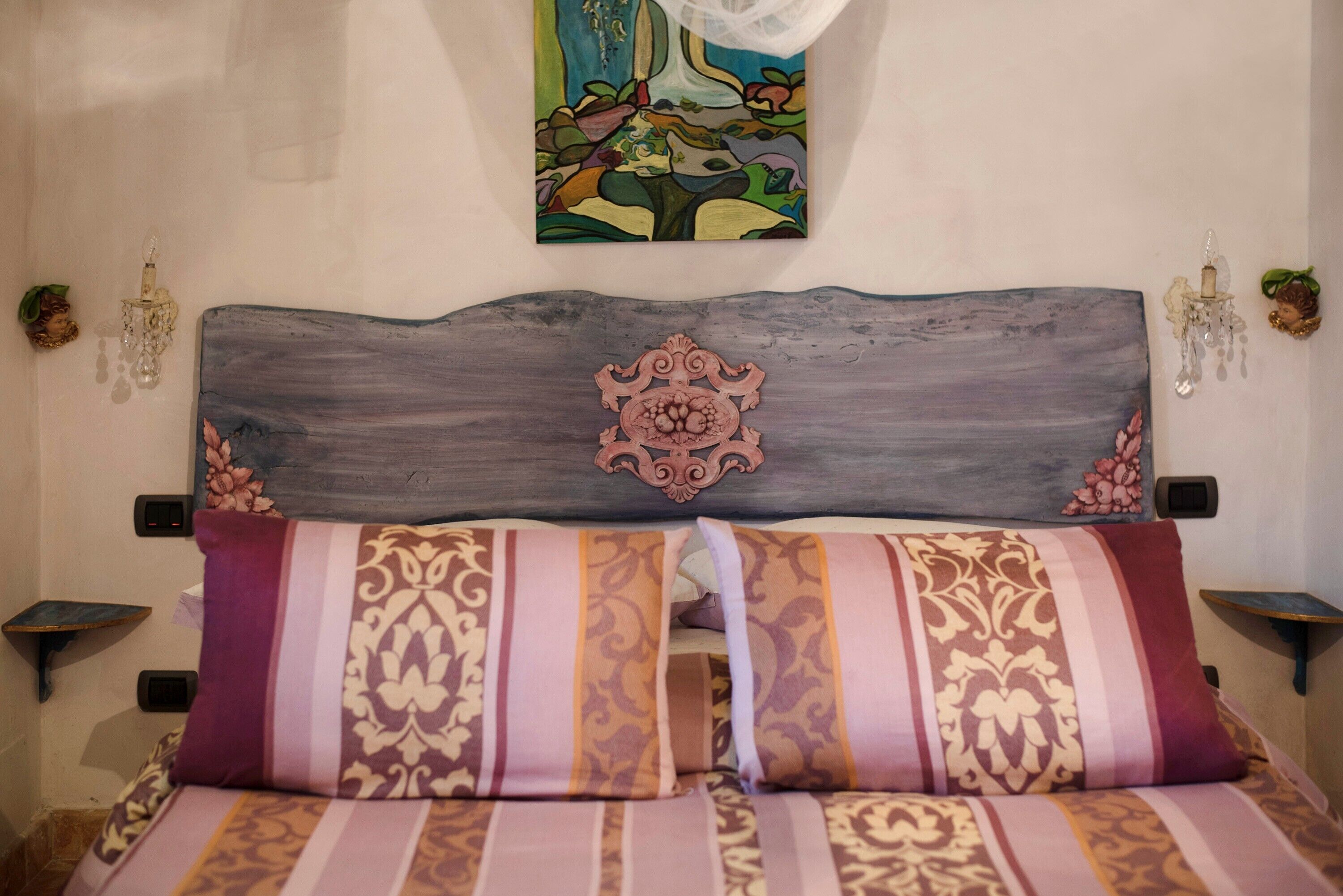 A bed with striped bedding, a decorative headboard, a wall painting, and wall-mounted light fixtures.