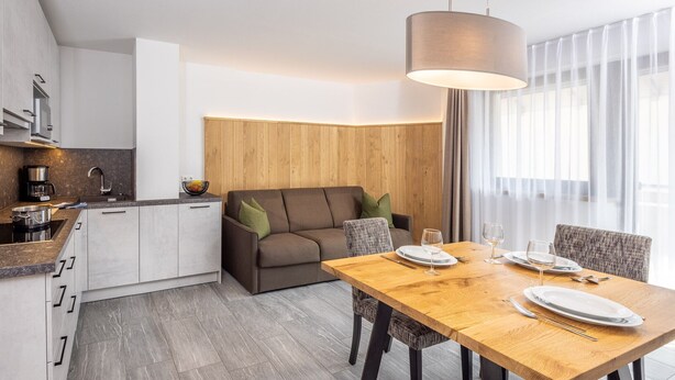 A modern kitchen with built-in appliances, a dining area with a wooden table and chairs, and a cozy seating area with a sofa and cushions. - Apartment 'Zum Sonnentor - Larix' with Mountain View, Balcony & Wi-Fi (Luttach)