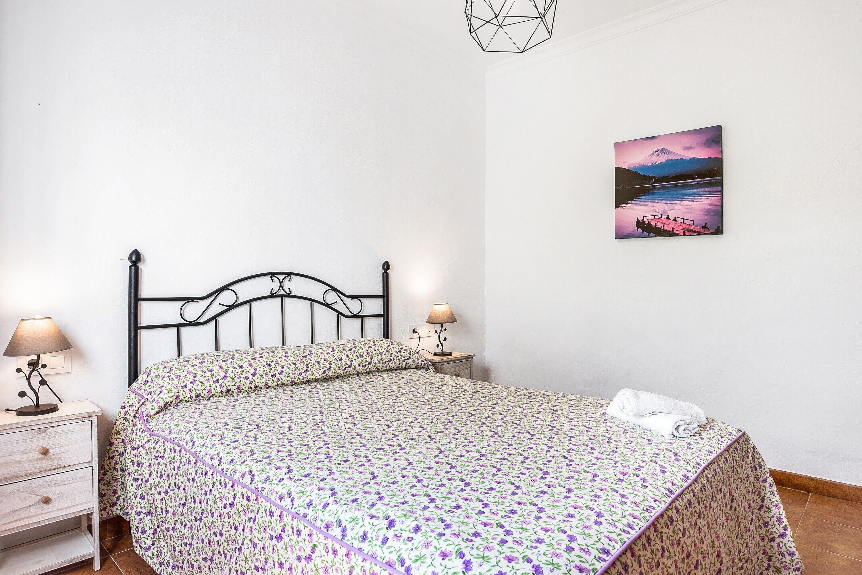 Een keurig opgemaakt bed met een bloemenbedekking, een nachtkastje met een lamp en een wanddecoratie van een berglandschap.