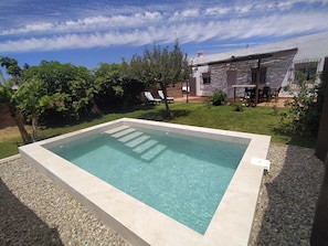 A rectangular swimming pool with clear water, surrounded by a pebble border, in a backyard with a house, lawn, and trees.