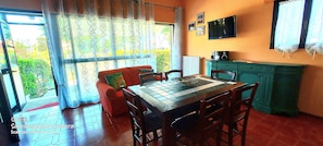 A dining area with a table and chairs, a sofa with cushions, a green cabinet, and a television on the wall. - Casa Europa 🏡 is your vacation home at 400mt. from the sea with private garden. (Cavallino treporti)