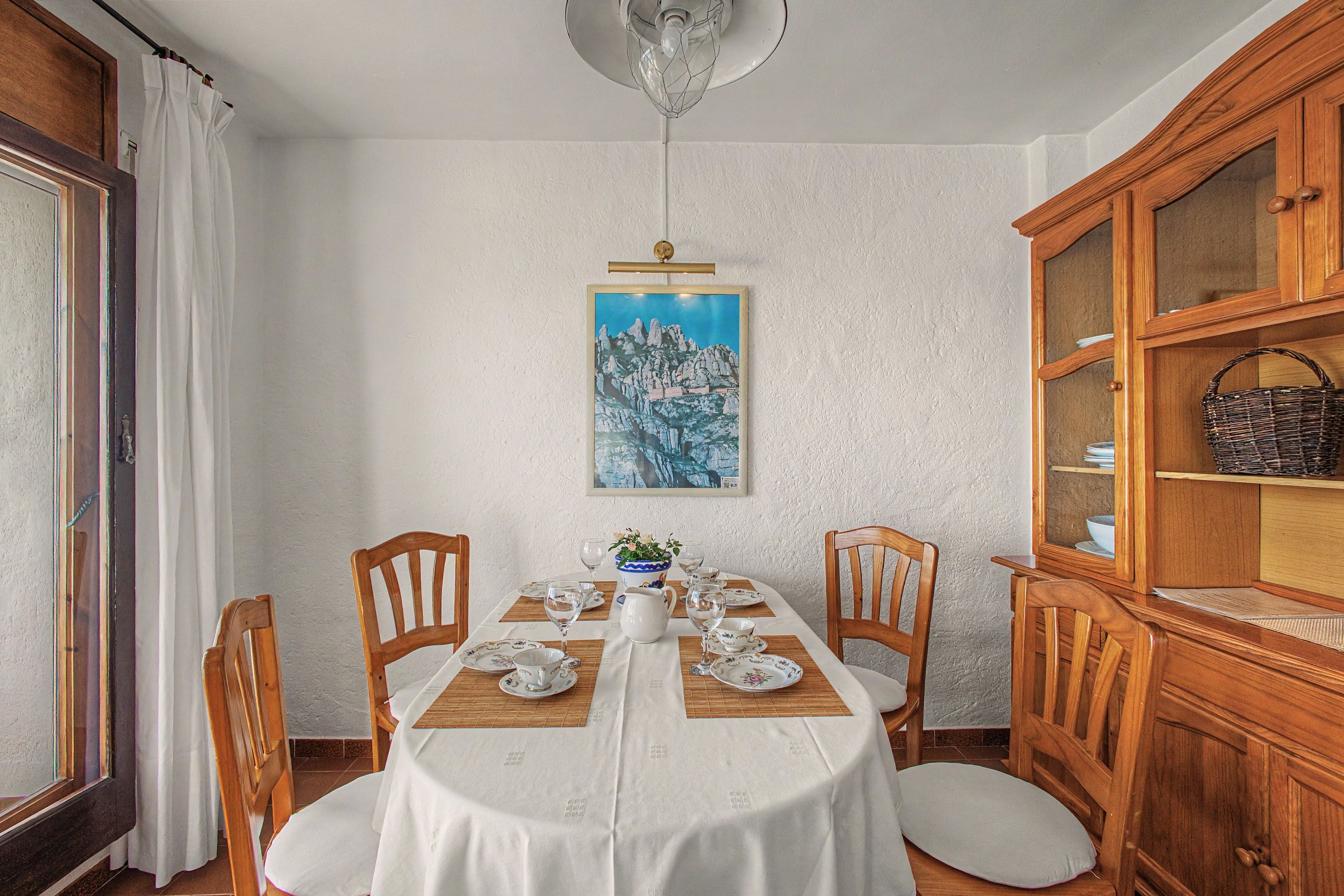 A dining room with a round table set for four, wooden chairs, a white tablecloth, a painting on the wall, and a wooden cabinet with glass doors.