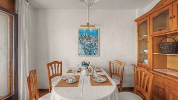 A dining room with a round table set for four, wooden chairs, a white tablecloth, a painting on the wall, and a wooden cabinet with glass doors.