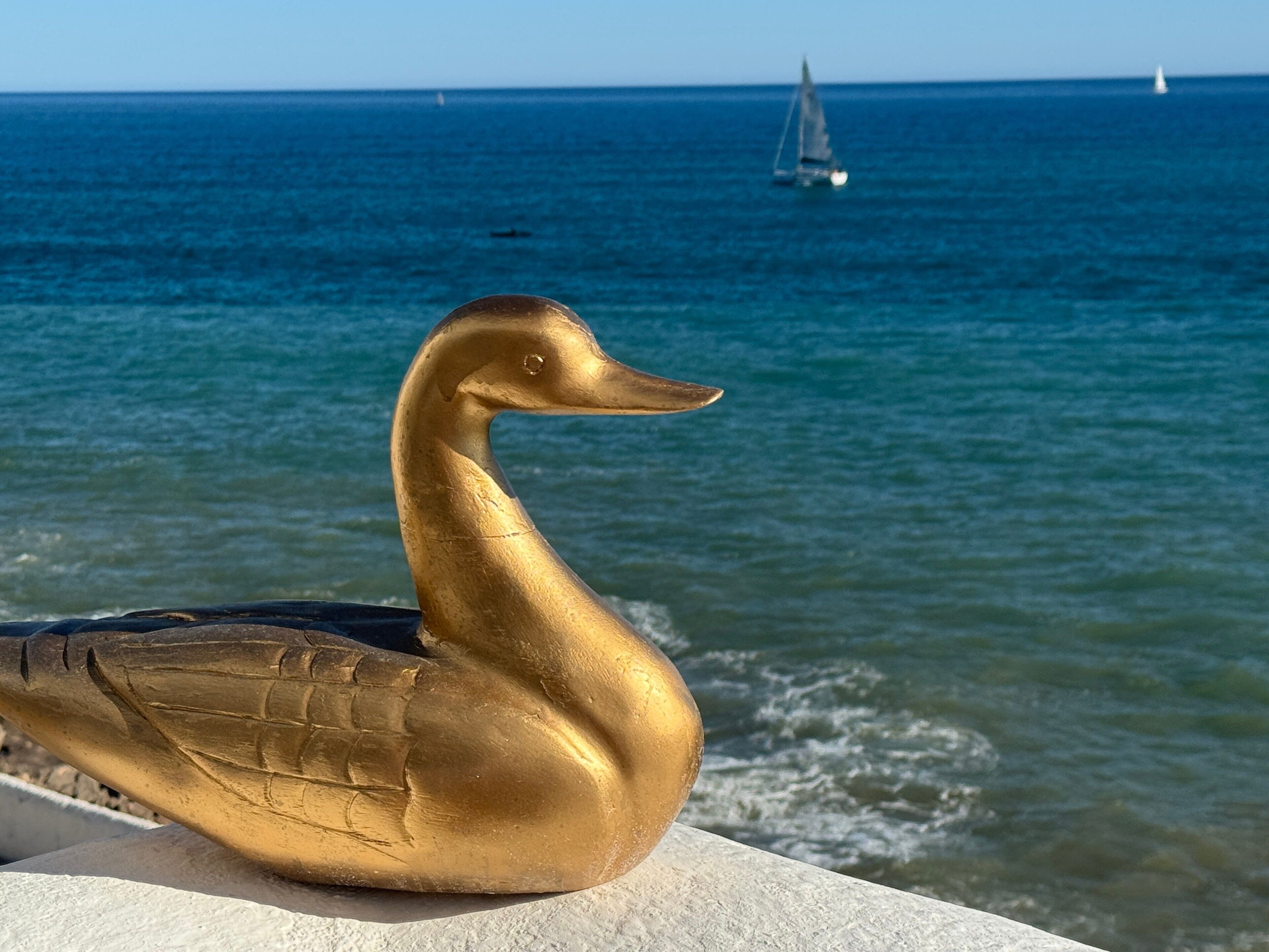 Une sculpture dorée représentant un oiseau au bord de la mer, avec un voilier en arrière-plan.
