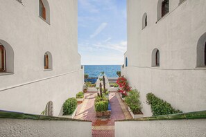 Ein Gang mit Blick auf das Meer, begrenzt von weißen Gebäuden mit Rundbogenfenstern, der zu einer mit Pflanzen bepflanzten, gefliesten Terrasse führt.