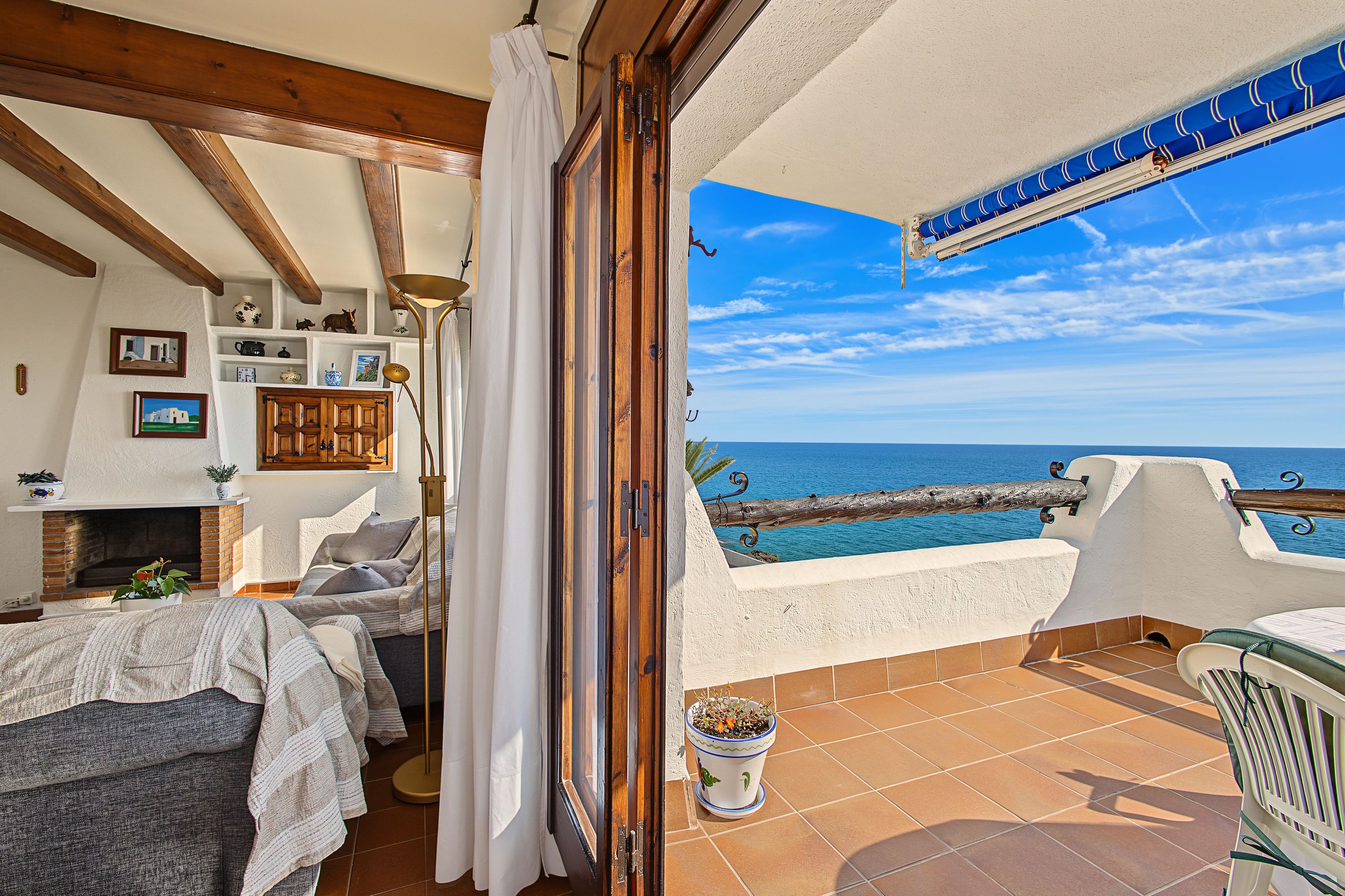 A room with a bed, a fireplace, a view of the sea, and a balcony with a chair and a potted plant.