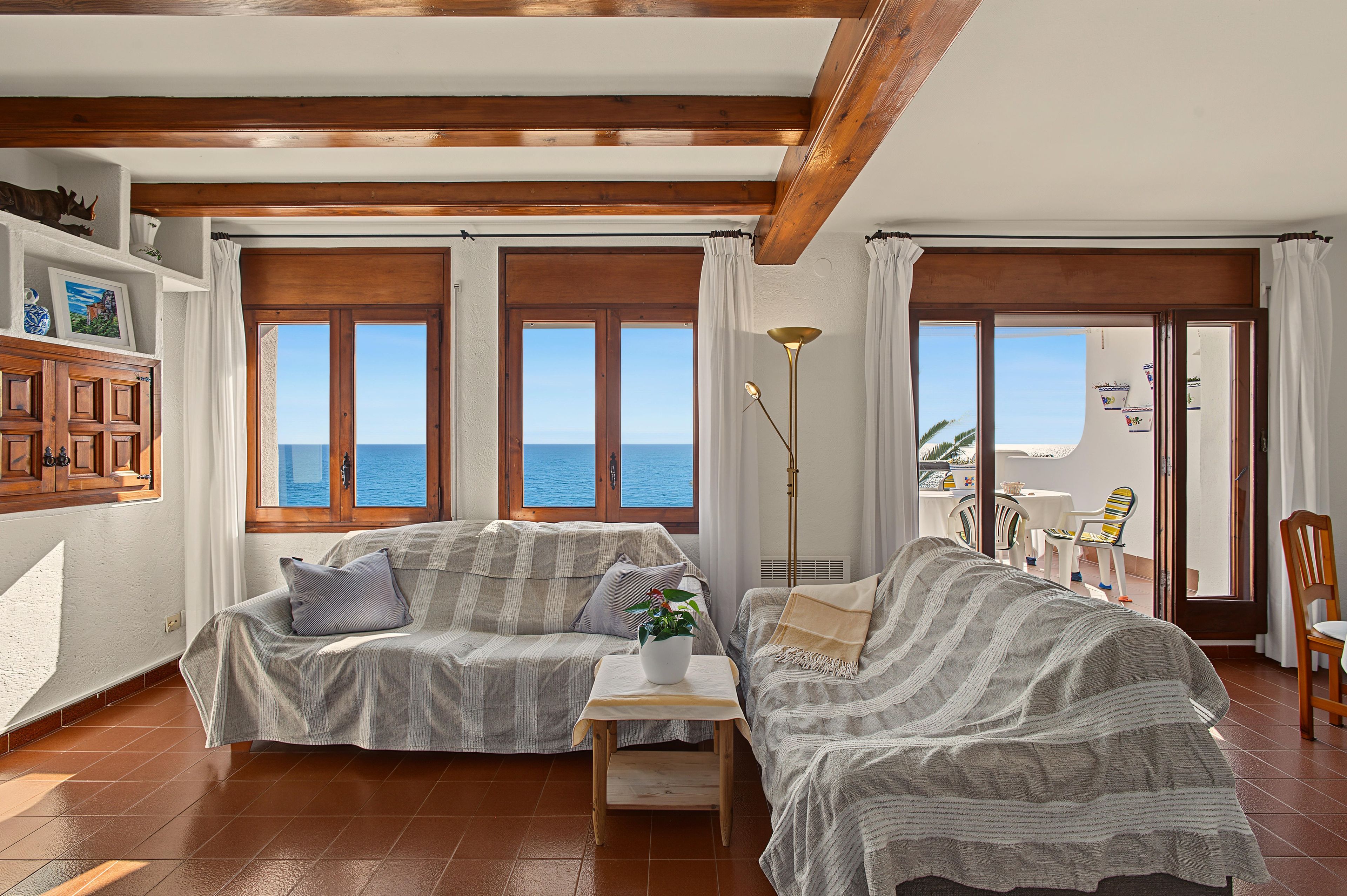 A cozy living room with a wooden ceiling, a sofa, a small table, and a view of the sea through large windows.