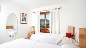 Et hotelværelse med to senge, et træpanel bag hovedgæret, et rundt spejl, et indrammet billede, et vindue med gardiner, en stol, et natbord med lampe og vase, samt udsigt til et bjerg.