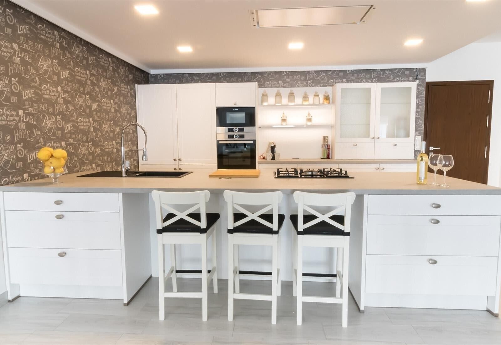 Una cocina moderna con armarios blancos, una isla central con taburetes altos, horno empotrado y salpicadero con azulejos decorativos.