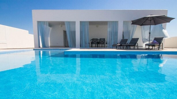 A clear blue pool with a modern white building in the background, featuring a covered outdoor seating area with umbrellas. - Casa Rural "Villa MyJ" with Pool, Garden, Terrace & Wi-Fi (La Puebla de Cazalla)