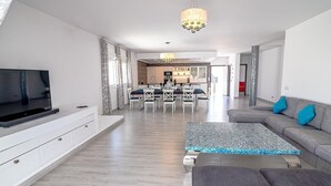 A modern living room with a flat-screen TV, grey sofa, patterned rug, and a dining area with a chandelier. - Casa Rural "Villa MyJ" with Pool, Garden, Terrace & Wi-Fi (La Puebla de Cazalla)