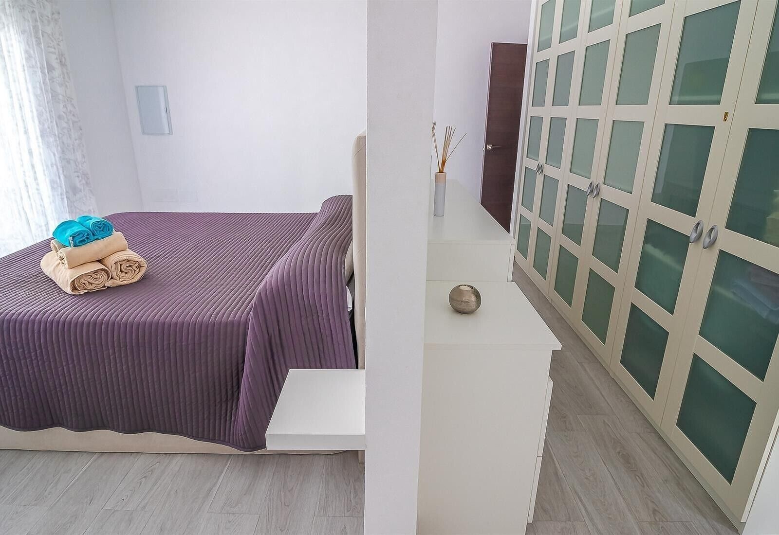 A room with a bed, a small table with a vase, and a wardrobe with glass doors.