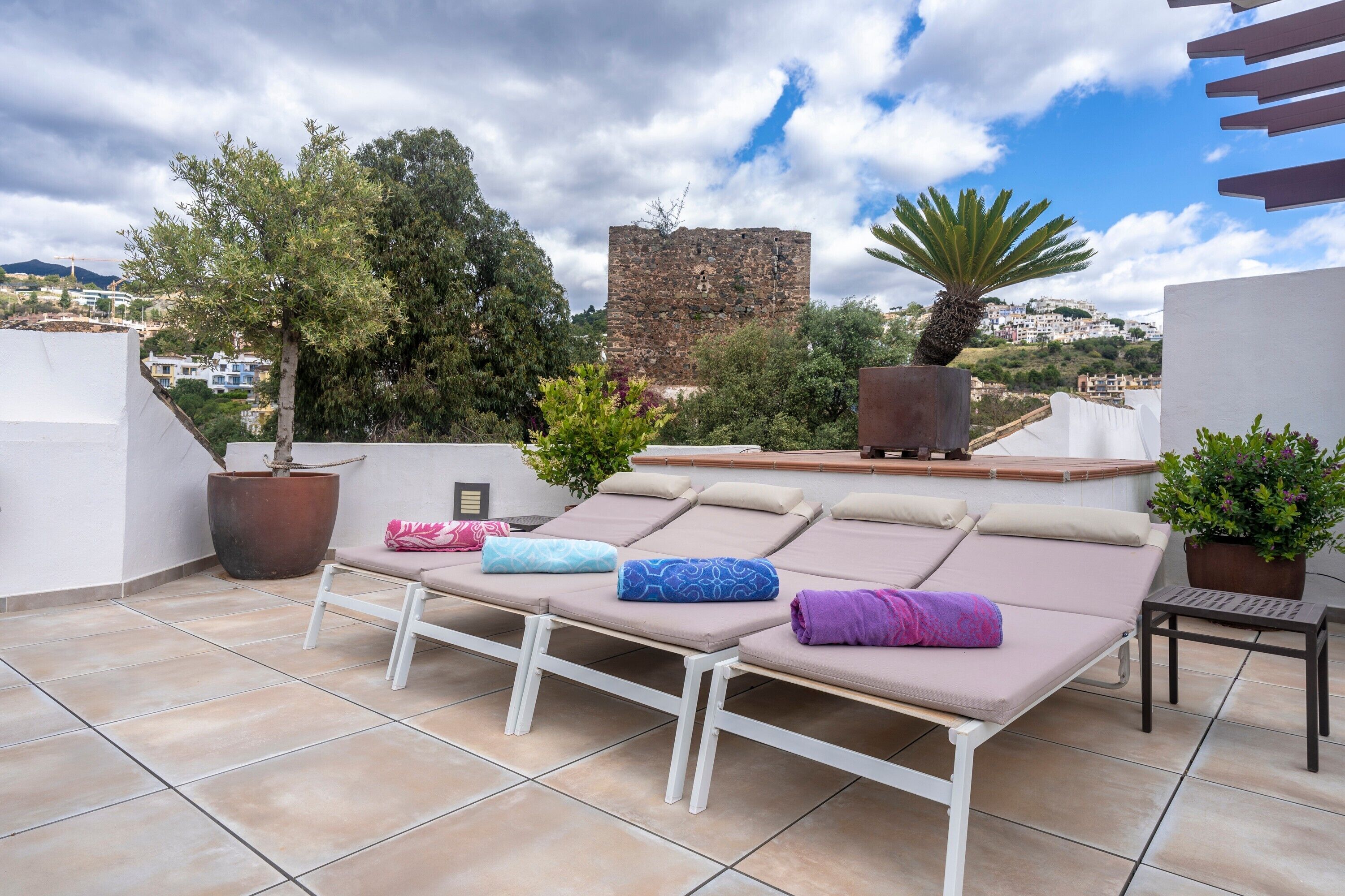Terraza en la azotea con sillones de descanso, plantas en macetas y vistas a una antigua torre de piedra.