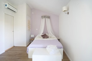A bedroom with a bed, white curtains, a bedside lamp, and a stack of towels.