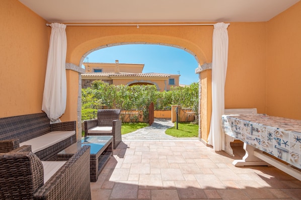 A sunlit patio with wicker furniture, a pool, and a view of a building through an archway.