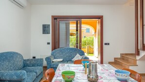 Una sala de estar con un sofá, sillas, una mesa con mantel estampado y vistas a un espacio exterior a través de un ventanal.