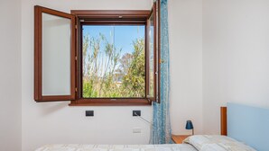 Un dormitorio con cama, ventana con vista a zonas verdes, mesita de noche con lámpara y toma de corriente.