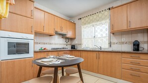 A kitchen with wooden cabinets, a round table with chairs, a built-in oven, and a window with patterned curtains. - Pet-friendly holiday home 'Casa Quintião' with Wi-Fi (Ferragudo)