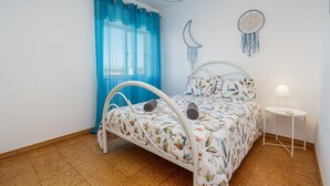 A bedroom with a bed, a small table, a lamp, a window with blue curtains, and wall decorations. - Pet-friendly holiday home 'Casa Quintião' with Wi-Fi (Ferragudo)