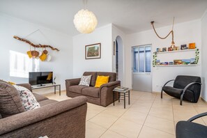 Een woonkamer met een bank, fauteuils, een televisie, een hanglamp en een boekenkast.