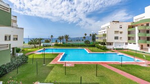 Zona de piscina con vista al mar, rodeada de vegetación y edificios.