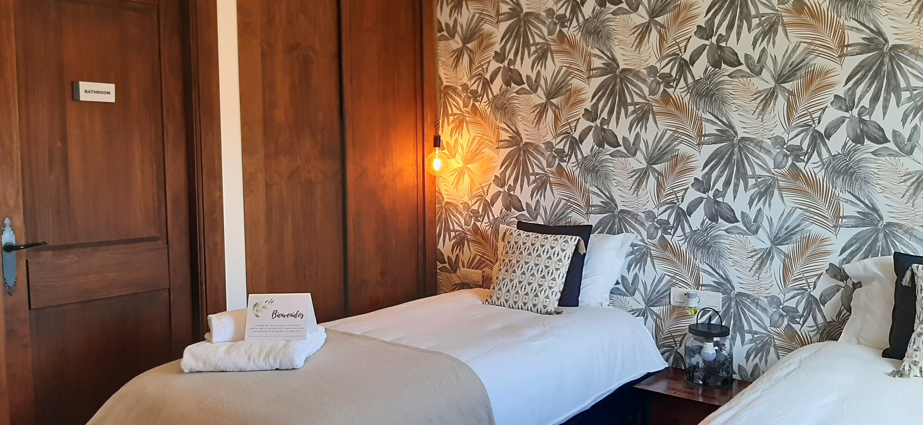 A hotel room with two beds, a wooden wardrobe, a tropical leaf-patterned wall, and a welcome card on the bed.