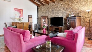 A living room with a stone wall, wooden beams, pink sofas, a flat-screen TV, and a fireplace. - Villa Elsa with Pool, Garden, Terrace & Wi-Fi (Conil de la Frontera)